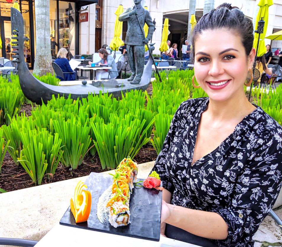 Yuly - smiling lady sitting in a restaurant patio hiding a plate of sushi